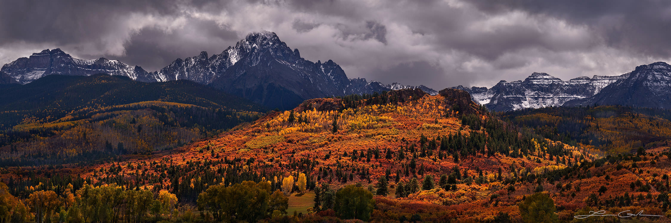 A colorful hill of orange and yellow fall colors, illuminated by light and surrounded by mountains