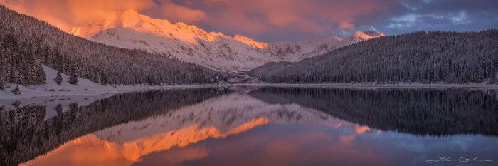 A calm lake reflecting snow covered trees and mountains in orange sunset light and clouds