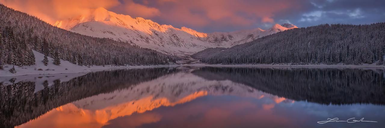 A calm lake reflecting snow covered trees and mountains in orange sunset light and clouds - Small