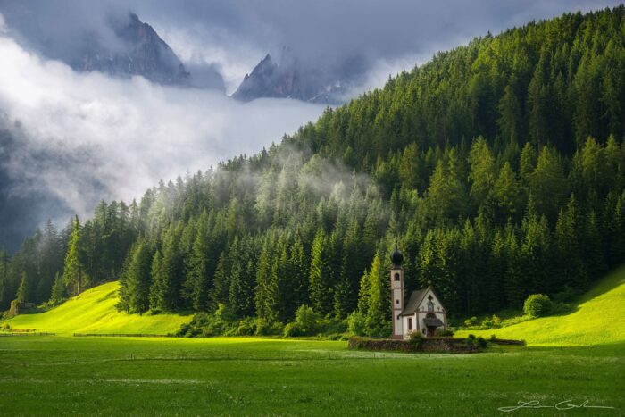 A beautiful tiny church on a green meadow at the foot of a forest and tall mountains with some fog and clouds above, Dolomites, Italy
