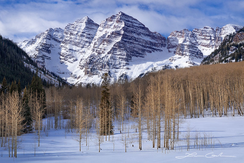 Jagged peaks of Maroon Bells with white clouds and blue skies, and an aspen forest during winter - Small