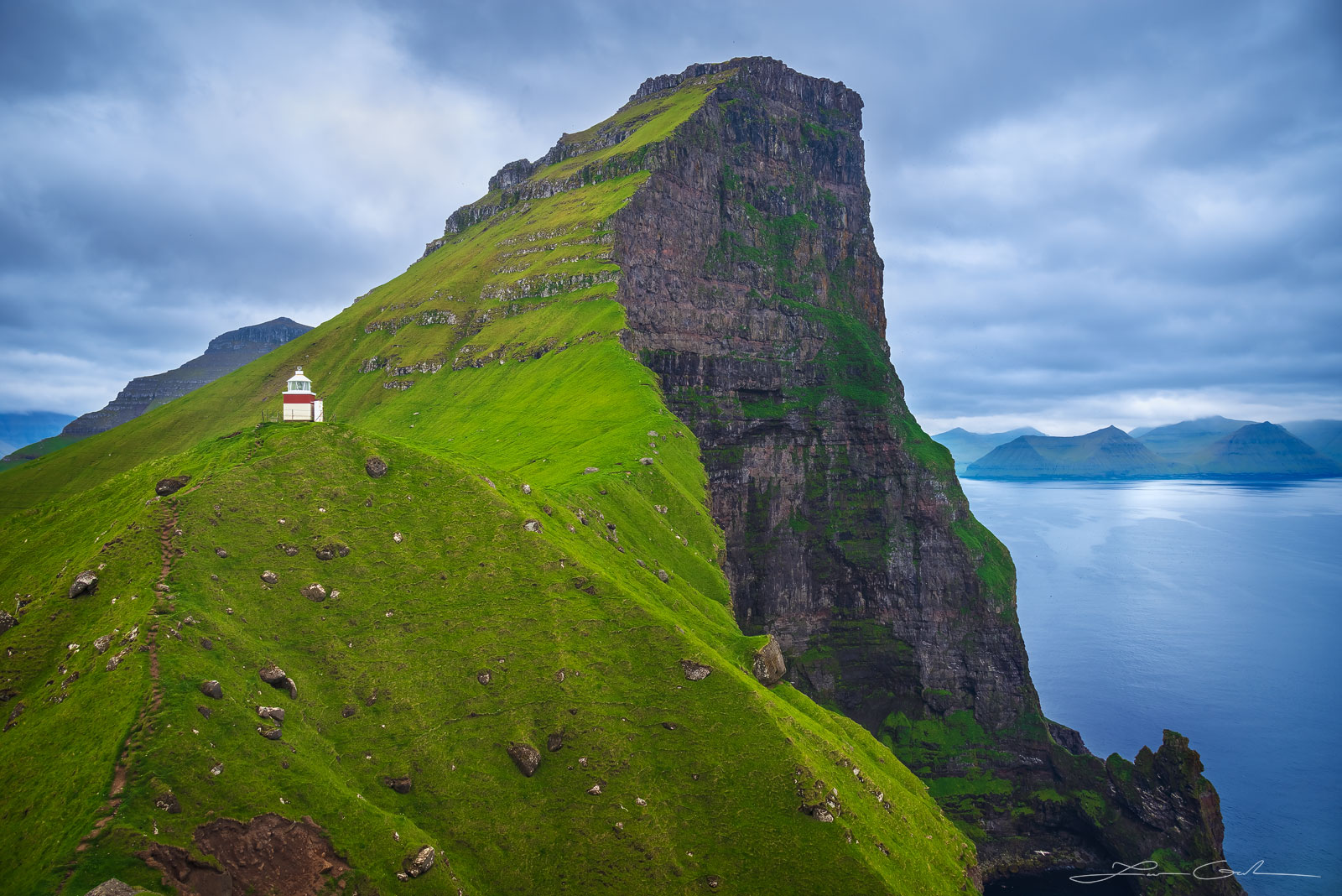 A fine art photograph of a lighthouse high above the ocean, next to a mountain with green grass - Faroe Islands - Gintchin Fine Art