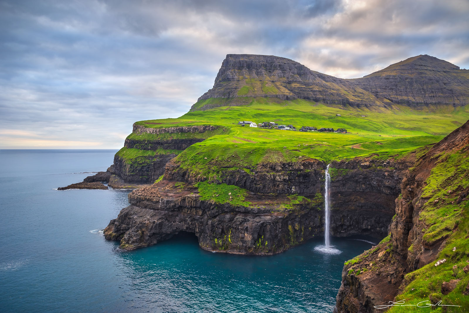 A beautiful waterfall flowing into the ocean from a green lush valley surrounded by mountains - Faroe Islands - Gintchin Fine Art