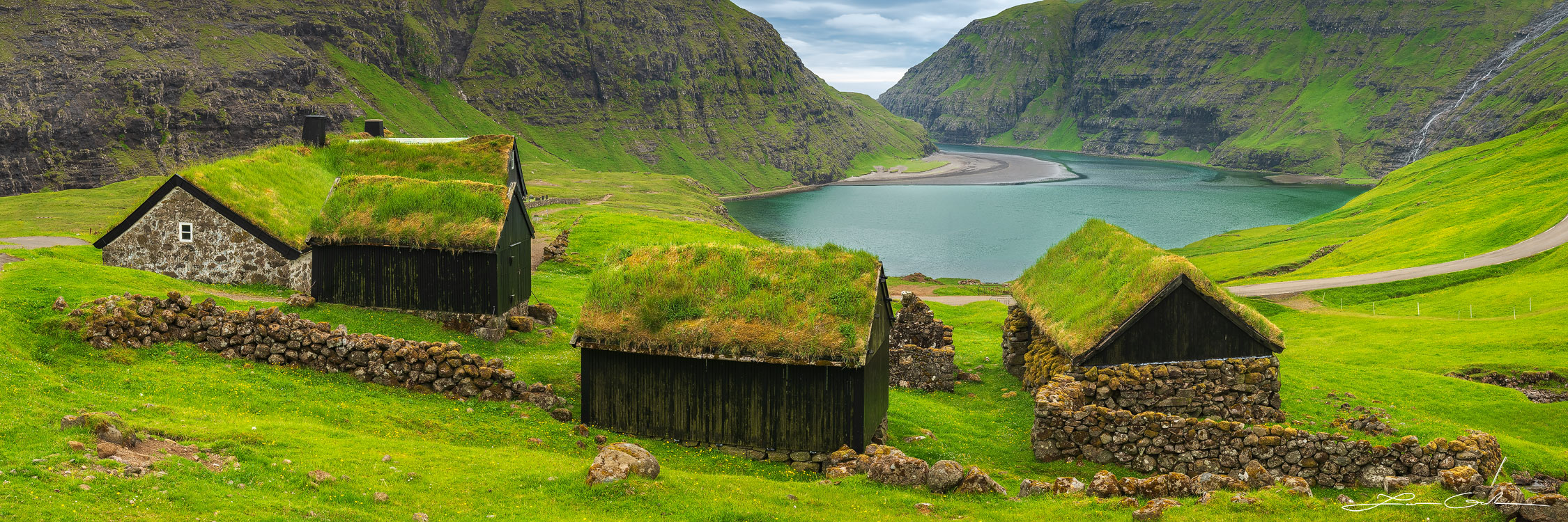 Grass roof houses surrounded by lush meados and mountains with a lake and a waterfall in the distance - Faroe Islands - Gintchin Fine Art