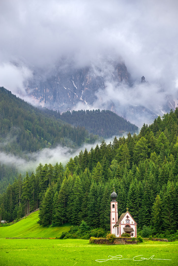 A small mountain church surrounded by lush green meadows, forests, and steep rock walls in South Tyrol, Italy - Gintchin Fine Art