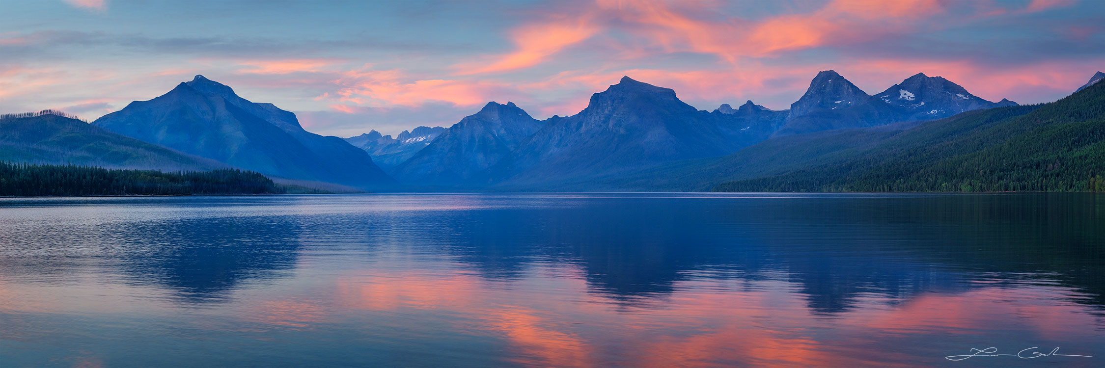 A panorama of a big mountain lake and lots of mountains in the distance, reflecting in the water at sunset with some pink and orange clouds - Glacier National Park, Montana - Gintchin Fine Art