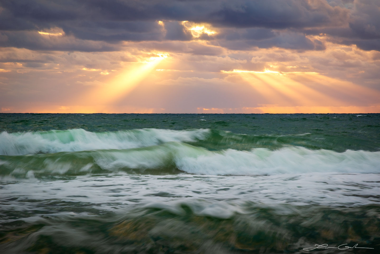 A wavy ocean with white foaming waves and dramatic sun rays peeking down through the clouds - Gintchin Fine Art