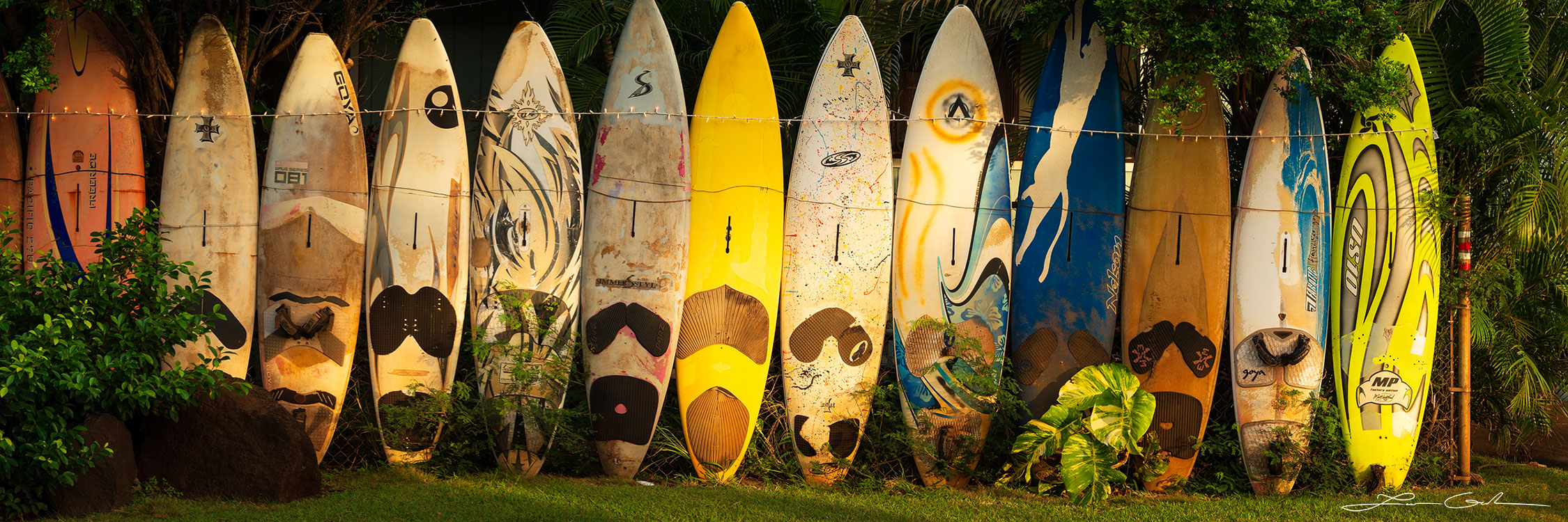 Panoramic photo of a row of vertical surfboards with diverse designs, serving as a fence and bathed in the glow of the rising sun, symbolizing the Hawaiian spirit - Gintchin Fine Art.