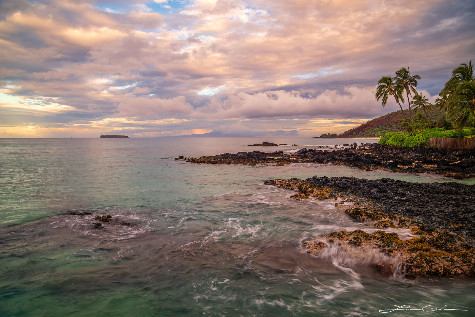 Sunrise over Maui coastline featuring soft, silken ocean waves against a volcanic shore, green and turquoise water, distant view of Molokini Island under light orange-hued morning clouds - Gintchin Fine Art.