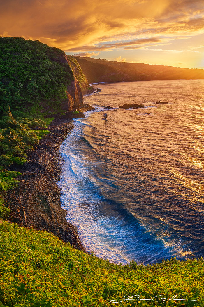 High-angle shot from cliff-top capturing a dramatic ocean sunrise over a pebbly shore, with lush cliff-side vegetation in Maui - Gintchin Fine Art.