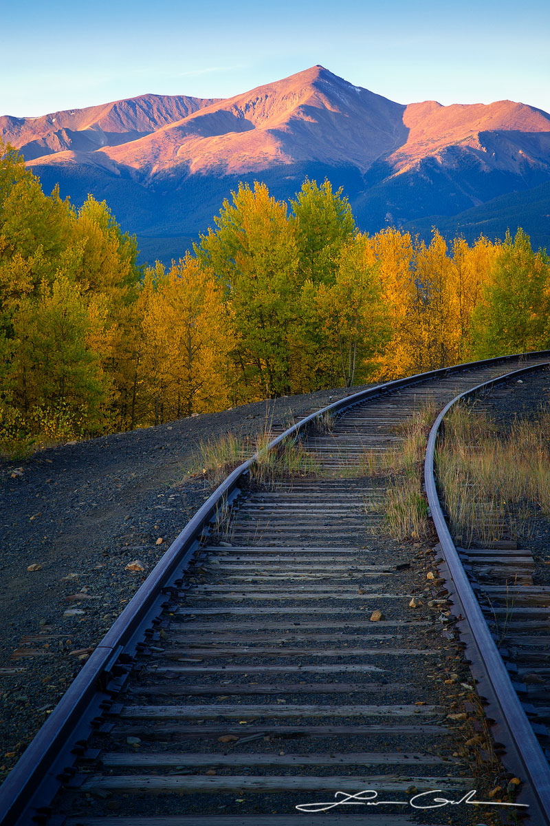 Scenic view of old abandoned mountain railroad track merging into distant aspen trees, with Mount Elbert in the backdrop against a clear blue sky - a unique portrayal of mountains, fall colors, and a railroad track. - Gintchin Fine Art