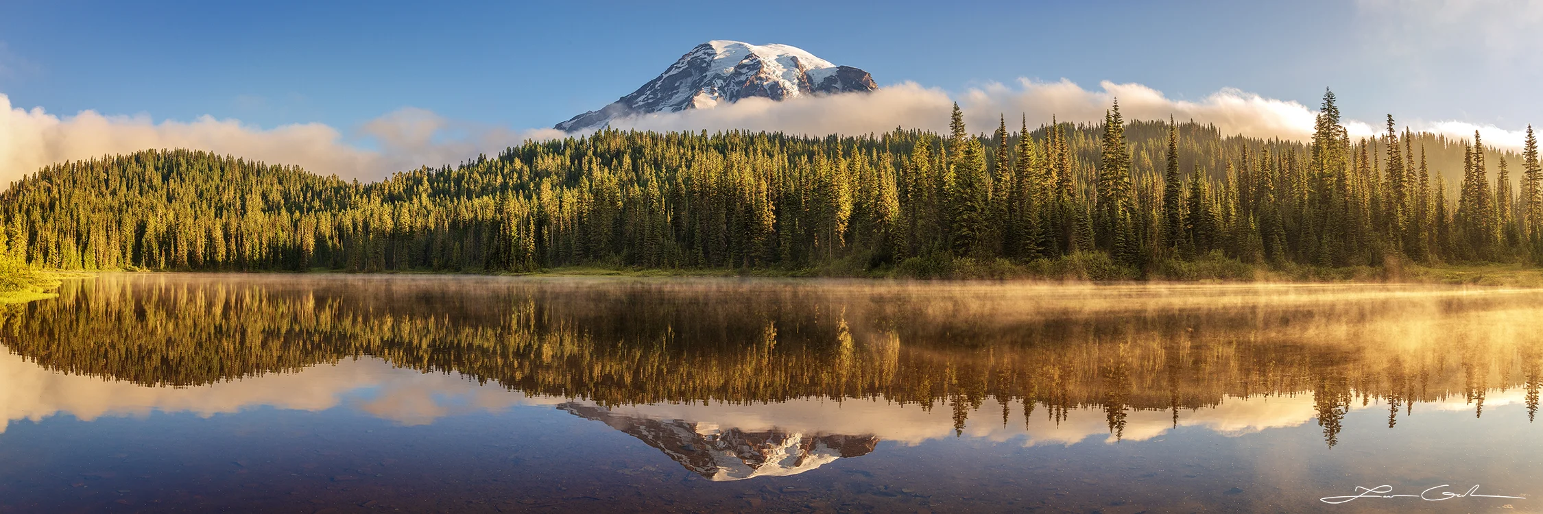 A tranquil photograph capturing the reflection of Mt Rainier in a calm lake at sunrise, with some lingering fog, a clear blue sky, and evergreen trees on the shore. - Gintchin Fine Art
