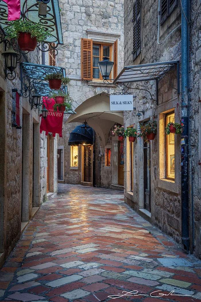 A photo print of a quaint European street in Kotor, Montenegro, featuring a narrow cobblestone alleyway lined with charming shops, vibrant flower pots, and rustic stone buildings with wooden shutters. - Gintchin Fine Art