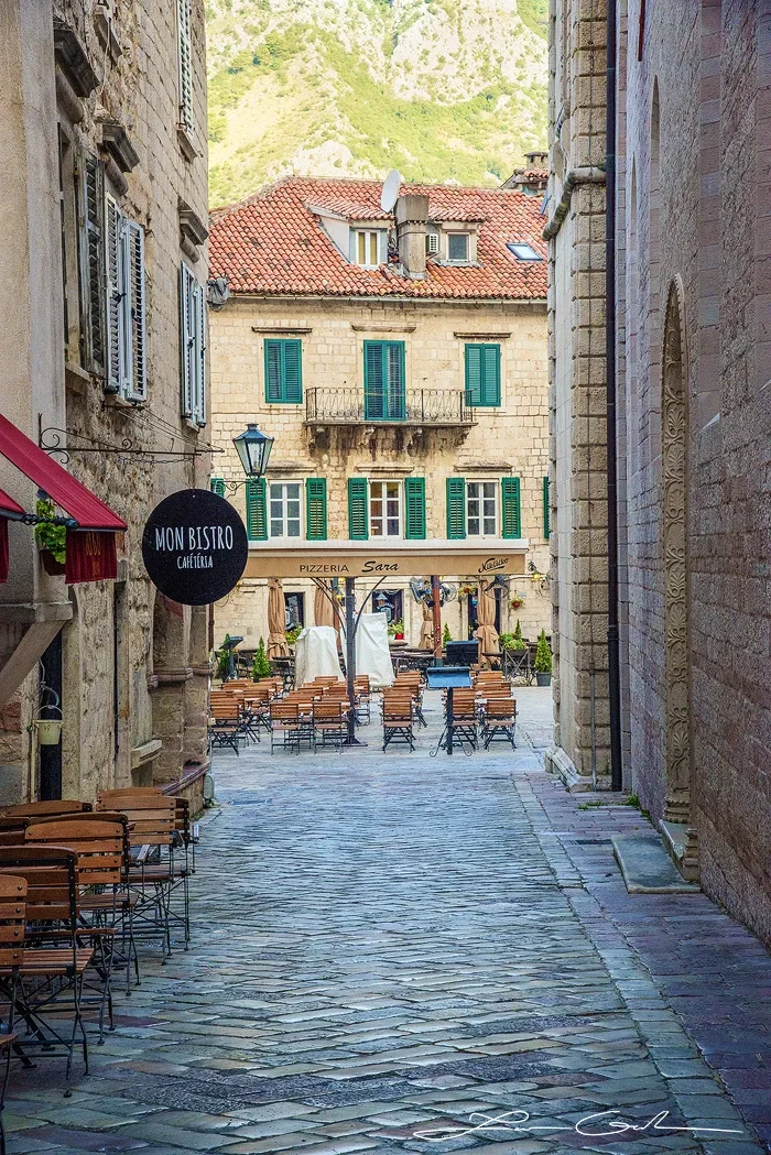 Fine art photo print of a European town square in Kotor, Montenegro, featuring a cobblestone alleyway, green shutters, red-tiled roofs, outdoor cafes, and distant mountains. - Gintchin Fine Art