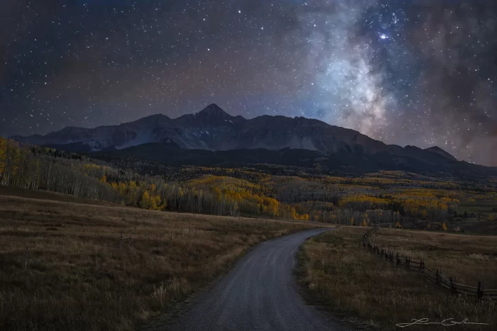 Nighttime landscape of Wilson Peak near Telluride, Colorado, with the Milky Way galaxy illuminating the sky. A winding dirt road leads through a field bordered by golden aspen trees, while Wilson Peak rises in the background against the starry night. The Milky Way stretches diagonally across the sky, creating a stunning contrast between the rugged mountain and the vast cosmos above.