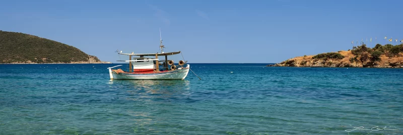 A traditional Mediterranean fishing boat floats in turquoise waters, surrounded by coastal hills under a clear blue sky. - Gintchin Fine Art