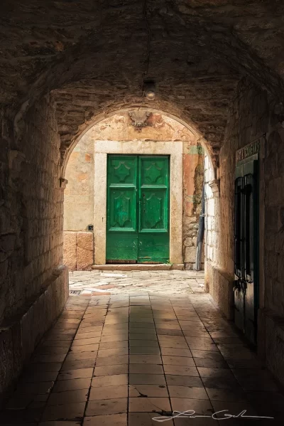 Fine art photo print of a historic passage in Kotor, Montenegro, featuring a green ancient door framed by a stone archway and a cobblestone path. - Gintchin Fine Art