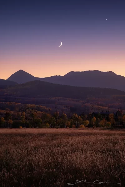 A serene twilight scene featuring a crescent new moon and the planet Venus aligned above the silhouette of Colorado mountains during autumn. The foreground showcases a golden-brown field and vibrant autumn foliage, with the majestic Rocky Mountains in the background, capturing the peaceful beauty of a Colorado autumn under the new moon.