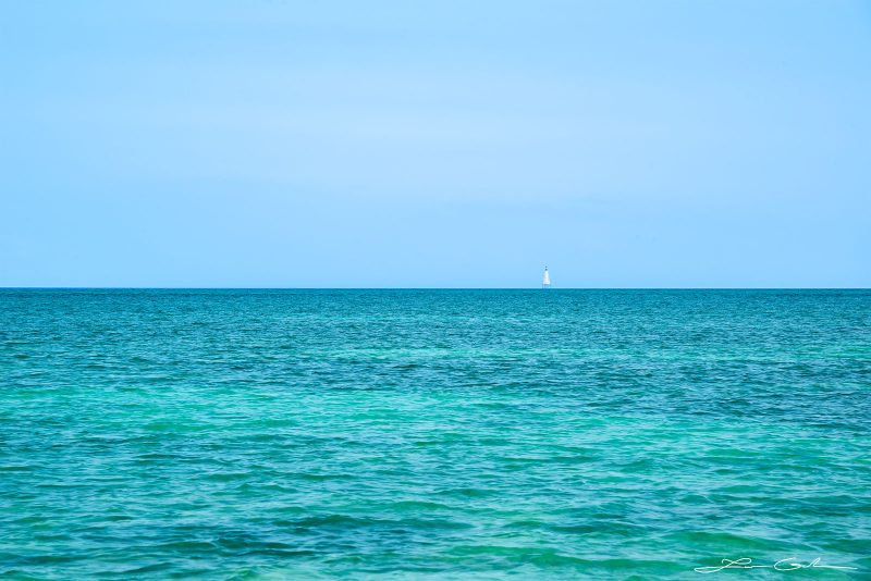 A minimalistic image of turquoise ocean, clear skies and a light house on the horizon in the distance - Gintchin Fine Art